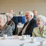 Two women laugh while seated at a table, with a man seated behind them out of focus